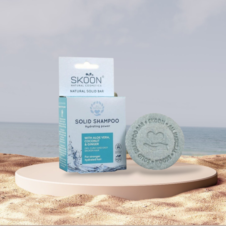 Pureday En Skoon solid shampoobar Hydraterende kraft og dens emballage vises på en rund sten mod en rolig strandbaggrund. Emballagen fremhæver naturlige ingredienser som aloe vera og ingefær, hvilket fremmer dens økologiske appel.