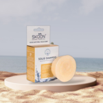 Pureday En Skoon shampoobar - Sensitive Moisture & Care ved siden af emballagen på en cylindrisk piedestal mod en strandbaggrund. Pakken fremhæver naturlige ingredienser som mandelmælk og avocado, med vægt på velvære.