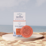 Pureday En Skoon Color & Shine naturlig kosmetik shampoobar og dens emballage, vist på et beige stativ mod en sandstrandbaggrund. Shampooen er cirkulær, orange og præget med mærkenavnet.