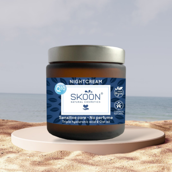 Pureday En krukke Økologisk natcreme - Skoon - Sensitive med q10 og naturlige kosmetiketiketter, sat mod en rolig strandbaggrund, placeret på en rund stensokkel. Produktet markedsføres til følsom hud uden parfume.