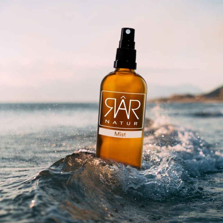 Pureday En sprayflaske af glas mærket "RAR Mist økologisk Rose hydrolat" er balanceret på en bølgetop ved stranden, med vandstænk omkring sig og en sløret baggrund i havet, der fremkalder en følelse af nat.