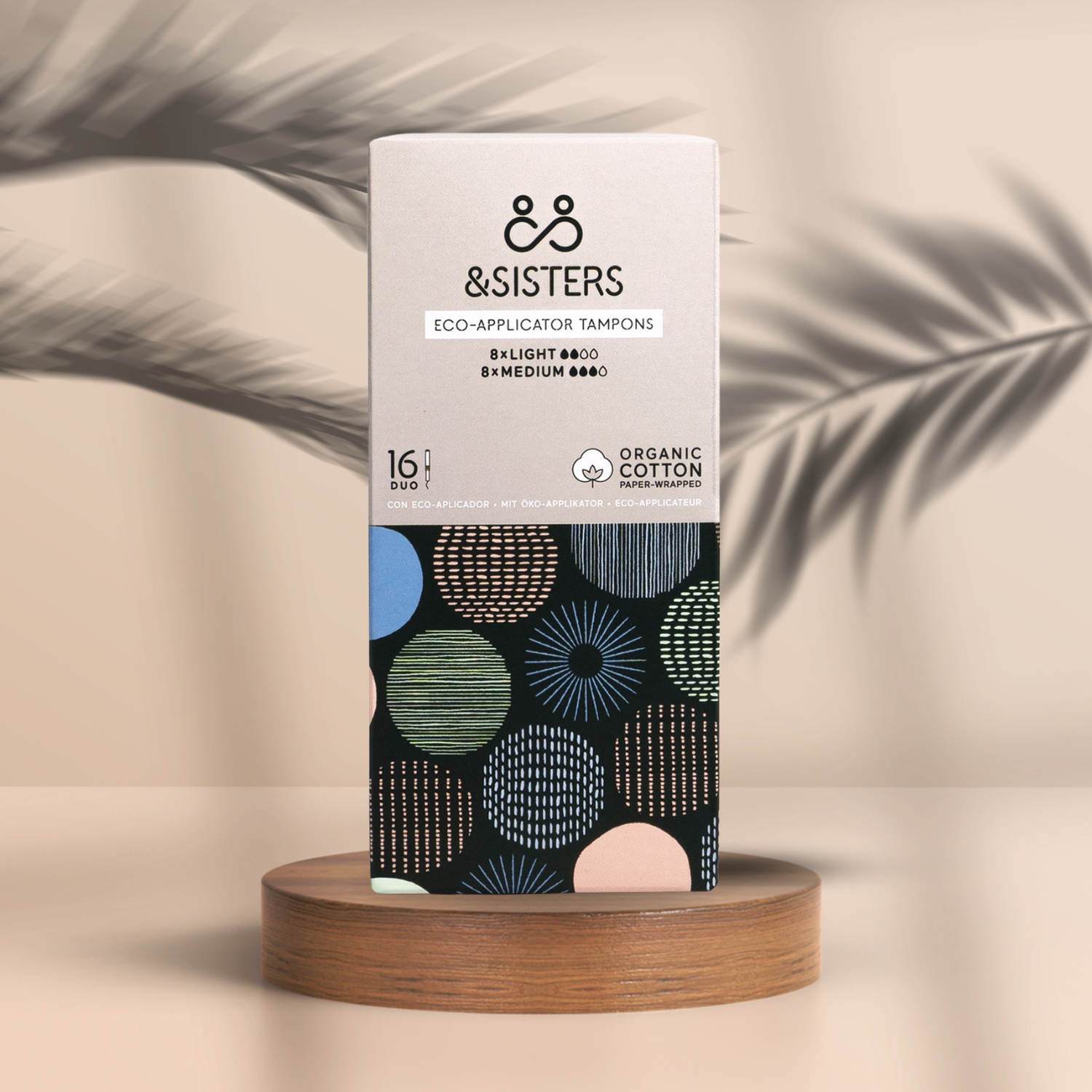 Pureday En æske Menstruation Tampon 8 Light & 8 Medium - Økologisk tampon fra &Sisters, designet til optimal velvære, hviler på et træstativ mod en blød beige baggrund, med et mønster af farverige cirkler.