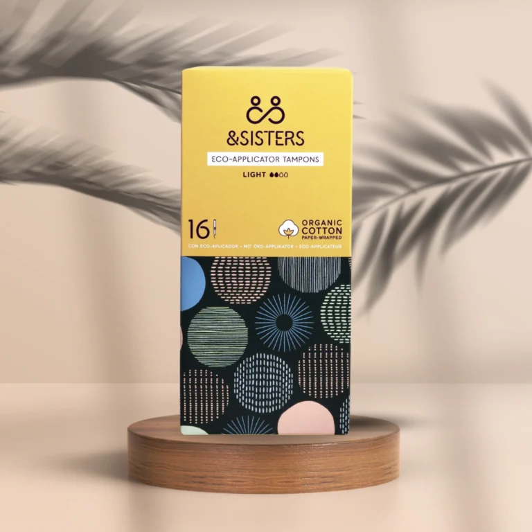 Pureday En æske Menstruation Tampon light - økologisk - &Sisters fra Matas står på et træstativ mod en beige baggrund; designet med gul emballage og et fedt, farverigt geometrisk mønster.