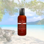 Pureday En flaske Skummende krops sæbe med citrongræs 375ml står på en sandstrand med klart blåt hav og frodigt grønt i baggrunden, og tilbyder en naturlig måde at vedligeholde velvære.