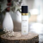 Pureday En flaske Gavesæt - Forkælelse til mor og baby hudpleje lotion står på en træskive, mærket som crafted with pureday økologi. Baggrunden har bløde fokusdetaljer med en hvid vase og lyse blomsterdekorationer.