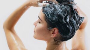 Pureday En kvinde med håret indskummet i Africa Organics - Mongongo Shampoo og balsam, hendes hænder løftet til hovedet, vasker hendes hår. Hun fremstår fredfyldt mod en almindelig, lys baggrund, der inkarnerer en følelse af velvære.